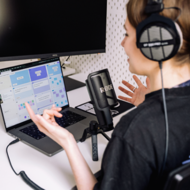 Eine Person mit Kopfhörern nimmt an einer Online-Schulung teil und spricht in ein professionelles Mikrofon. Auf dem Laptopbildschirm ist ein digitales Whiteboard mit Karten und Notizen zu sehen.